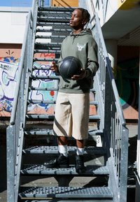 Ung mand på metaltrapper, iført en grøn hættetrøje med et logo, beige shorts og sorte sneakers, mens han holder en basketball.