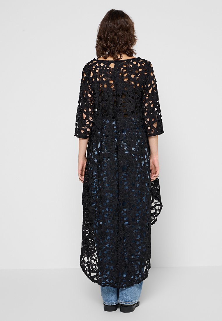 Schwarze Spitzen-Cardigan mit langen Ärmeln, offenem Design mit floralen Mustern und asymmetrischem Saum, getragen über blauen Jeans.