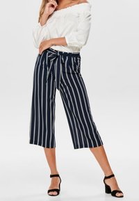 Kvinna klädd i marinblå och vita vertikala ränder avkortade byxor med knytmidja, en vit off-shoulder-blus och svarta klacksandaler.