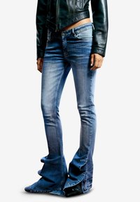 Pantalones vaqueros de mezclilla azul con bajo acampanado, con detalles de volantes alrededor de los tobillos. Textura desgastada con desvanecimiento ligero y costuras.