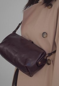 Mörkbrun läderväska med rektangulär form, med dragkedjestängning och avtagbar rem, bärs över en beige trenchcoat.