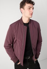 Maroon bomber jacket with a ribbed collar, zip closure, and side pockets. Worn over a black textured sweater; features quilted design details.
