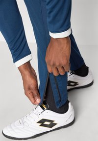 Σκούρο μπλε αθλητικό παντελόνι με λευκό περιλαίμιο, διακοσμημένο με φερμουάρ στο τελείωμα. Χέρι που προσαρμόζει το φερμουάρ· λευκά και μαύρα παπούτσια με ορατό λογότυπο.