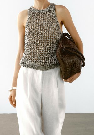 Personne portant un haut en maille sans manches et un pantalon blanc, tenant un grand sac tressé marron sous un bras sur un fond uni.