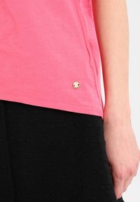 Lyserød bomulds T-shirt med korte ærmer, der har en rund kant og et lille metallisk logo på siden, parret med sorte strukturerede shorts.