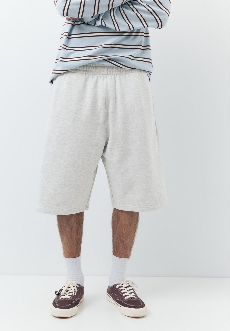 Mand iført lysegrå knælange shorts, hvide crew-sokker, vinrøde lærredssneakers og en blå og hvid stribet langærmet skjorte.