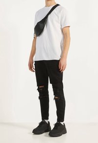 Mężczyzna w białej koszuli z krótkim rękawem, czarnych podartych dżinsach, czarnych sneakersach i czarnej torbie na ramię na jednolitym białym tle.