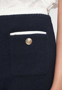 Gros plan sur la poche d'une jupe texturée bleu marine avec un bord blanc et un bouton doré, portée avec un top en dentelle blanche.
