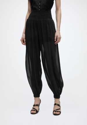 Vrouw draagt doorschijnende zwarte losse broek met elastische tailleband en boorden, gecombineerd met zwarte hoge hak sandalen met gouden details.