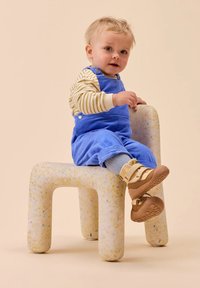 Des salopettes en velours côtelé bleu, une chemise à rayures à manches longues, des chaussettes grises et des chaussures beiges sur un enfant assis sur un tabouret blanc moucheté.