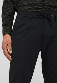 Pánské černé kalhoty s poutky na opasek a zavazovací šňůrkou v pase, v kombinaci s tmavým vzorovaným tričkem s dlouhým rukávem, ruka položená na boku.