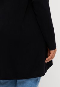 Personne portant un haut à manches longues noir et un jean en denim bleu, main détendue le long du corps, sur un fond clair uni.