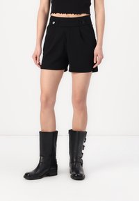 Shorts noirs ajustés avec deux boutons à l'avant, poches latérales et une texture de tissu lisse. Associés à de grandes bottes en cuir noires avec des boucles.