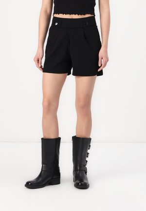 Shorts noirs ajustés avec deux boutons à l'avant, poches latérales et une texture de tissu lisse. Associés à de grandes bottes en cuir noires avec des boucles.