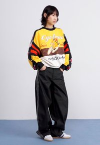 Kleurrijke oversized sweatshirt met grafische print in geel, rood en zwart, gecombineerd met losse zwarte broeken en witte sneakers.