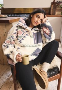 Pile con motivo geometrico multicolore, base crema e tasca blu. Modello seduto, che tiene una tazza verde, indossando pantaloni neri.