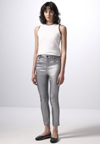 Stříbrné metalické slim-fit kalhoty s předními kapsami, doplněné o bílý top bez rukávů a černé boty s hroty. Hladký povrch a lesklý vzhled.