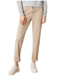 Osoba v béžových kalhotách s manžetami, šedém dlouhém rukávu a krémových teniskách stojící s rukama u těla.