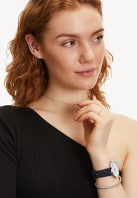 Zilveren sieraden met in elkaar verstrengelde cirkels op een fijne ketting, gedragen over een zwarte geribbelde top; een horloge met een marineblauwe band maakt de look compleet.