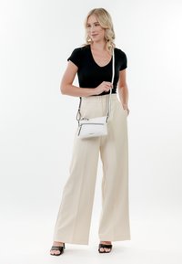 Top corto nero a maniche corte, pantaloni a gamba larga color crema e borsa a tracolla bianca con cerniere. Modella che indossa tacchi aperti neri, in piedi in un ambiente neutro.