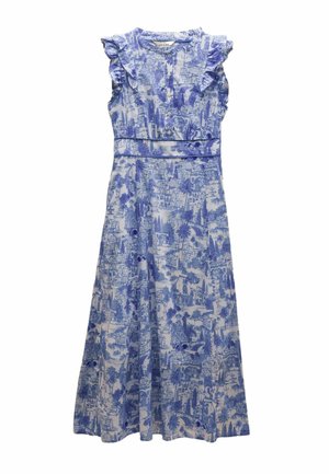 Kobieta stojąca w bezrękawniku, długiej sukience maxi w niebiesko-biały wzór z falbanami na ramionach, w beżowych klapkach, na tle beżowej ściany.