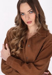 Bruine suède blouse met een kraag, voorzien van drukknoopsluitingen en een ontspannen pasvorm. De textuur is glad met een subtiele glans.