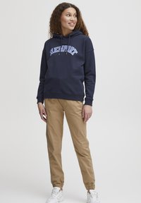 Felpa blu marina con il testo bianco "NEWPORT", vestibilità ampia, tessuto misto cotone, abbaiata con pantaloni chinos color beige, polsini elasticizzati e scarpe da ginnastica bianche.