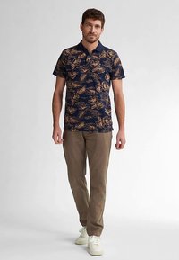 Marineblauw polohemd met crèmekleurig bloemenpatroon, korte mouwen en klassieke kraag, gecombineerd met lichtbruine chino's en witte sneakers.