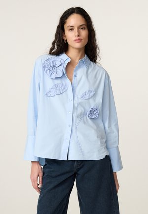 Vrouw draagt een lichtblauw overhemd met knopen en 3D bloem- en bladdetails, gecombineerd met donkerblauwe jeans tegen een effen achtergrond.