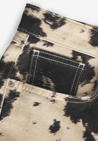 Denim beige délavé avec un motif tie-dye noir. Comprend une poche arrière avec des coutures blanches et un bouton en métal près de la ceinture.