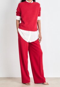 Pull tricot rouge à col rond, porté sur une chemise blanche à boutons. Un pantalon rouge à jambes larges et des sandales beige complètent la tenue.