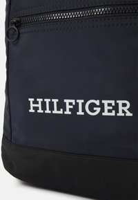 Sac à dos bleu marine avec des accents noirs, arborant un logo « HILFIGER » blanc bien en vue et un compartiment zippé. Texture lisse. Matériau durable.