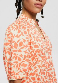 Blumenbluse mit cremefarbenem Grund und orangefarbenen Blumenmustern, einem V-Ausschnitt und kurzen Puffärmeln. Hergestellt aus leichtem Stoff.