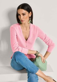 Rosa V-Ausschnitt-Cardigan, leichtes Strickmaterial, lange Ärmel. Getragen über blauen Jeans. Mit einer grünen gesteppte Handtasche und auffälligen Ohrringen.