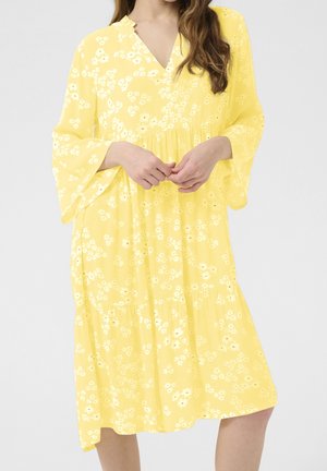 Woman in yellow V-neck dress with white floral pattern and wide sleeves, hands gently clasped at waist, standing against plain background.