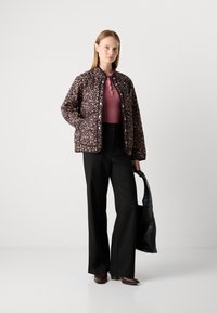 Woman wearing a floral quilted jacket over a pink ribbed top, black wide-leg trousers, brown shoes, holding a black handbag.