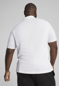 Polo shirt bianca a maniche corte esposta da dietro, con una vestibilità relax e tessuto di cotone morbido, dotata di un colletto classico e un design semplice.