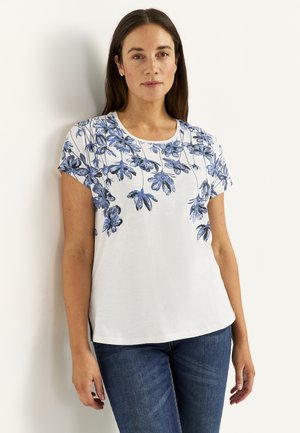 Frau trägt ein weißes kurzärmeliges Oberteil mit blauer Blumenmuster entlang des Ausschnitts, kombiniert mit blauen Jeans, steht vor einer hellen Wand.