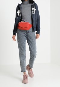 Riñonera de imitación piel roja llevada sobre una sudadera con cremallera azul marino y un cuello de tortuga gris. Jeans de tiro alto en un tono azul claro y zapatillas color rosa completan el conjunto.