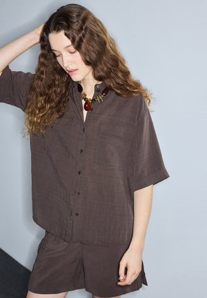 Junge Frau mit langen lockigen Haaren, die ein lockeres braunes Hemd mit Knöpfen und passende Shorts trägt, ergänzt durch eine auffällige Bernsteinkette.