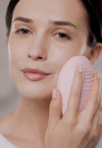 Silikon ansiktsrensende enhet, rosa, oval form med spor, holdt nær en persons ansikt. Glatt hudtekstur og naturlig leppefarge synlig.