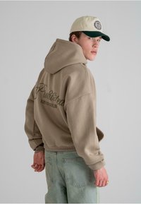 Beige hettegenser med hette og stor brodert tekst på ryggen. Kombinert med lyseblå jeans. Capen har et fargevalg i krem og mørkegrønn.