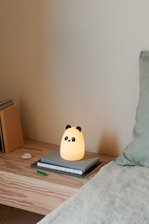 Lámpara en forma de panda con un suave cuerpo blanco luminoso y acentos negros. Colocada sobre libros apilados en una superficie de madera al lado de una cama.