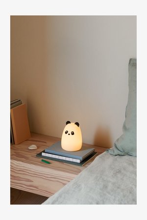 Lampa w kształcie pandy z miękkim, świecącym białym ciałem i czarnymi akcentami. Umieszczona na stosie książek na drewnianej powierzchni obok łóżka.