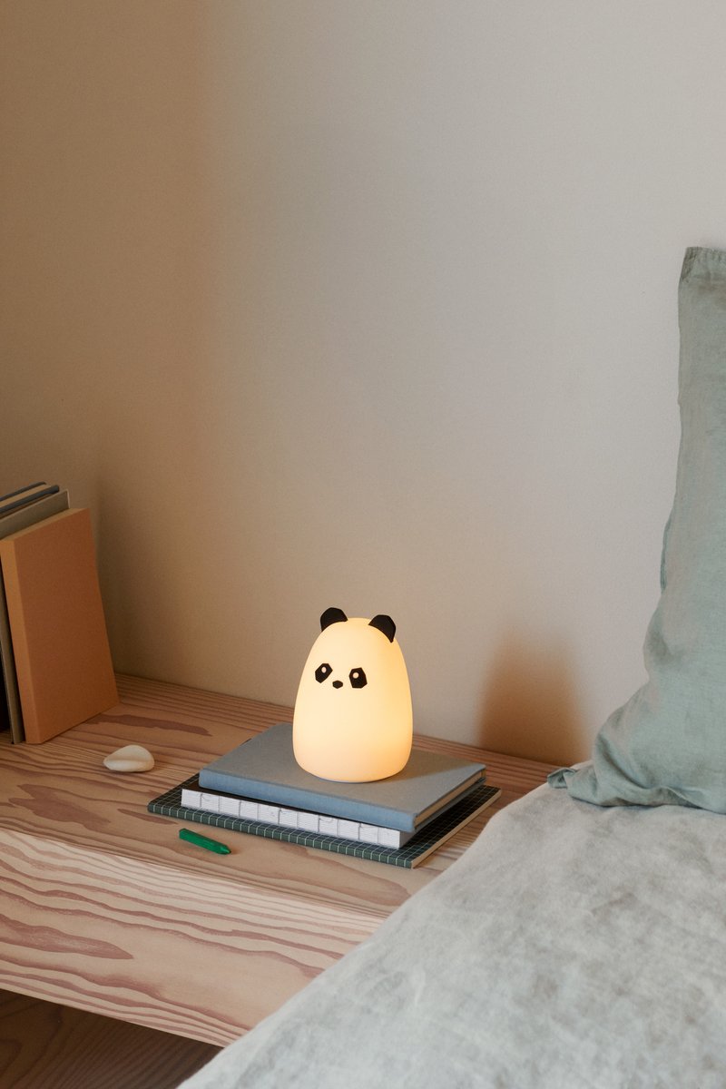Lampa v obliki panda z mehko, svetlečo belo telo in črnimi detajli. Postavljena na zloženih knjigah na lesenem površju ob postelji.