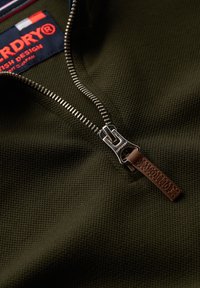 Tela texturizada de color verde oscuro con un diseño de media cremallera; cremallera de metal con una etiqueta de cuero marrón que dice "Superdry". Marca de contraste en el cuello.