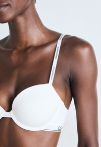 Close-up van een persoon die een witte Calvin Klein beha met merkbanden draagt tegen een lichte achtergrond.