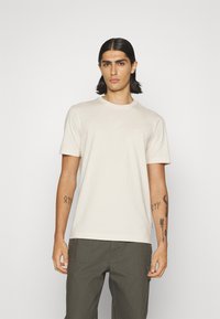 T-shirt en coton beige avec un col rond et des manches courtes. Détail de logo discret sur la poitrine ; modèle portant un pantalon vert olive.
