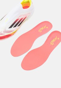 Scarpa da calcio bianca con strisce rosse e nere, caratterizzata da una parte superiore testurizzata e dettagli gialli. Due solette rosa sono posizionate adiacenti.