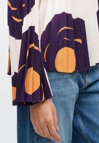 Színes, pliszírozott felső, krémszínű alapra nagy lila és narancssárga virágmintákkal, széles harang ujjakkal és texturált felülettel.